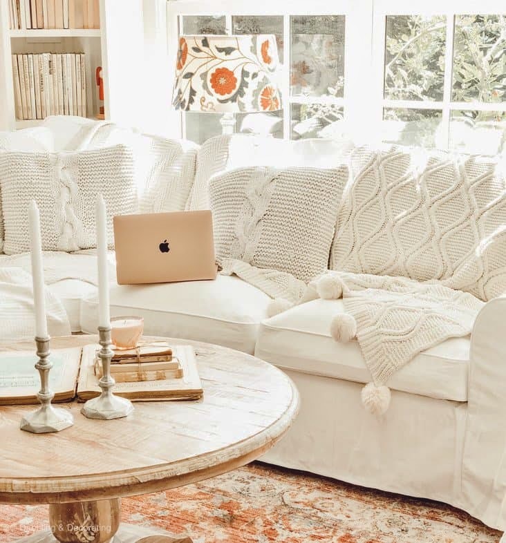 Cozy fall sunroom with white sectional, round wood coffee table and laptop open on layered blankets with white pillows.