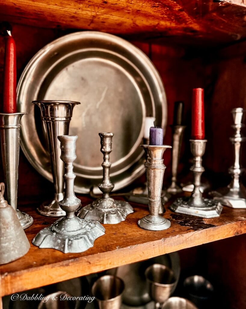 A collection of antique pewter candlesticks with different colored candles in antique hutch styling.