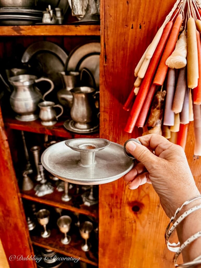Pewter candlestick in hand with styled antique hutch filled with pewter collection and hanging candles on door.