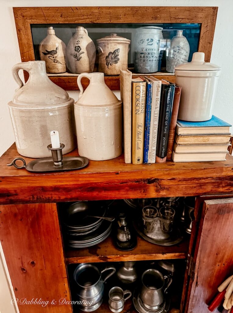 Top of antique hutch balanced and decorated with vintage crock jugs, vintage books and a pewter candlestick with a Goodwill framed picture on wall.