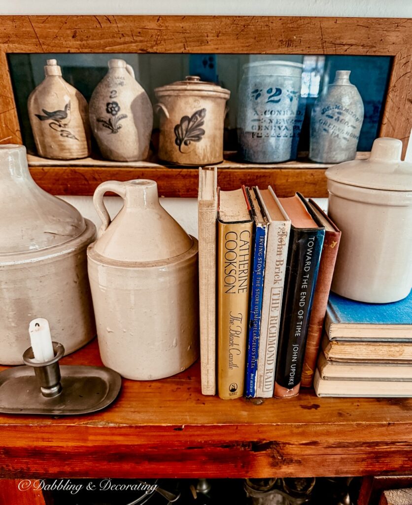 Top of vintage hutch with antique jugs, vintage books and pewter candlestick and framed wall picture of antique crocks.