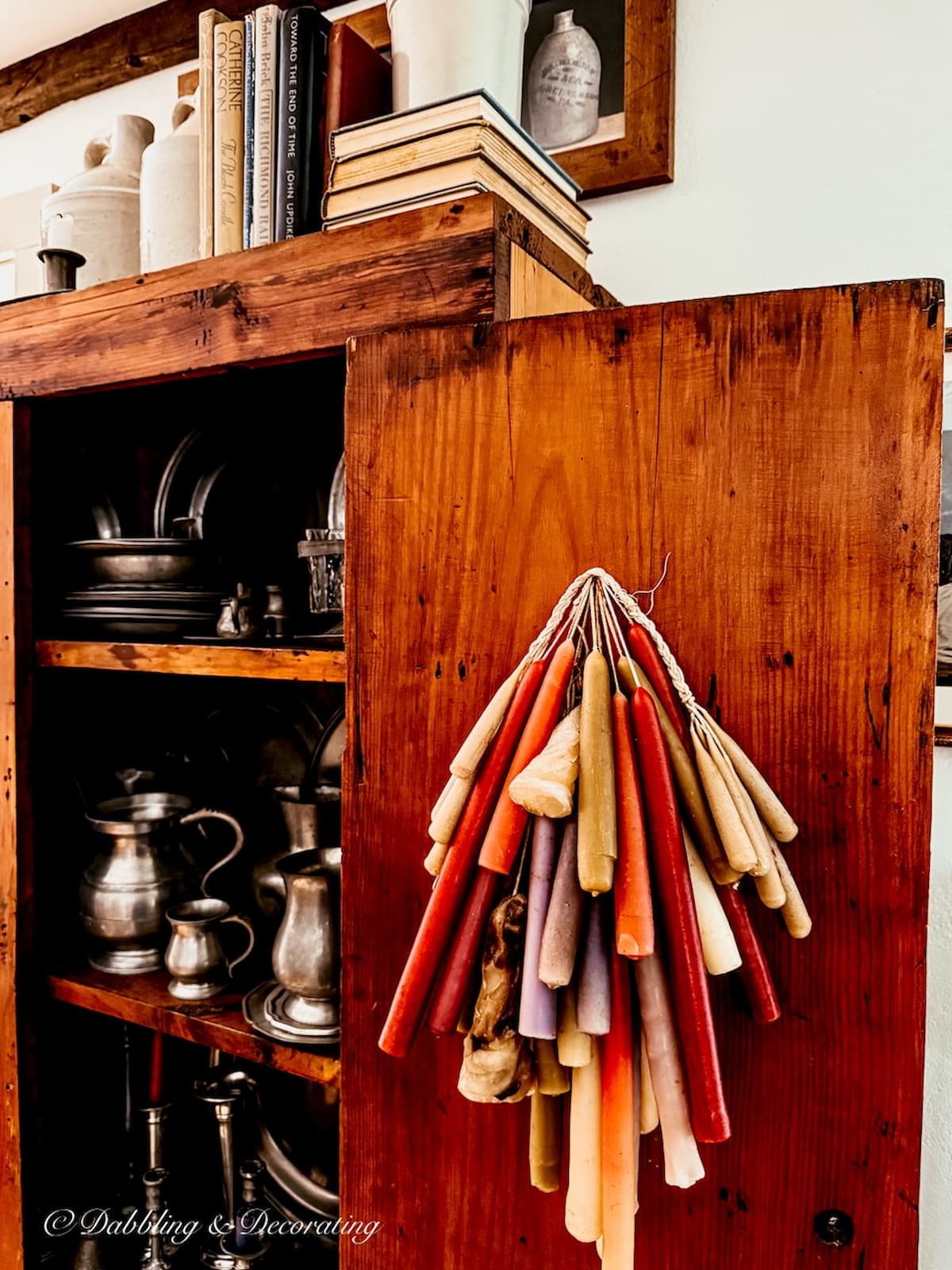 Antique hutch with pewter collection and a door filled with colorful candles in Early American home.