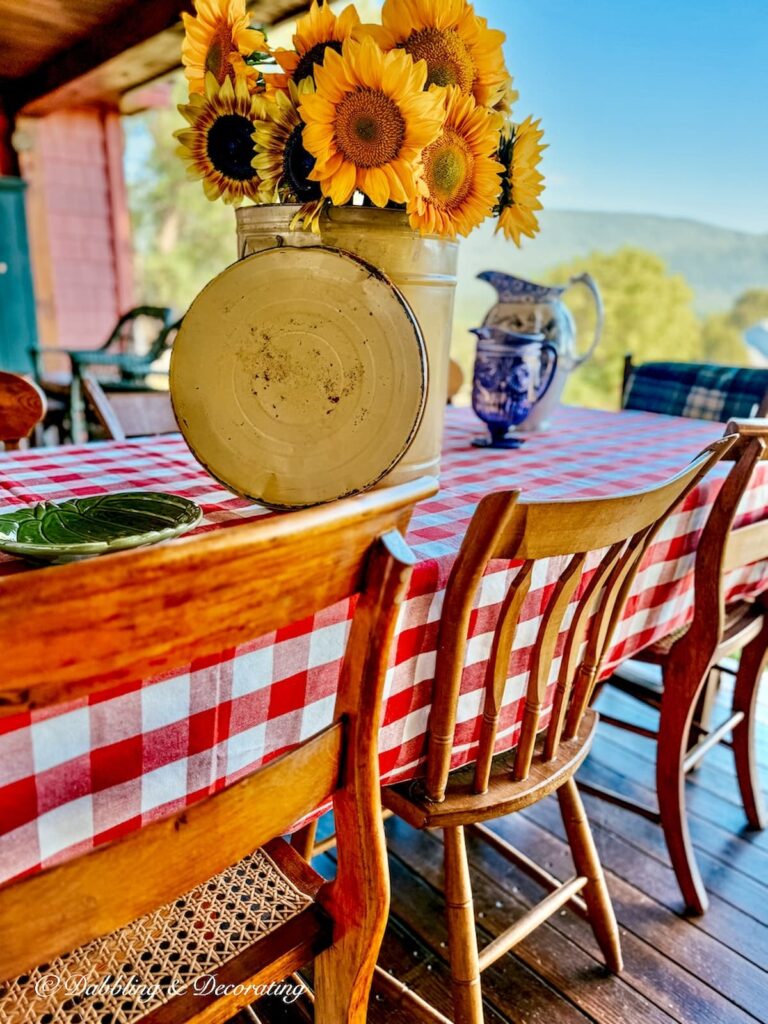 Yellow large vintage tin filled with sunflowers on outdoor porch table with red gingham tablecloth and vintage wooden chairs.