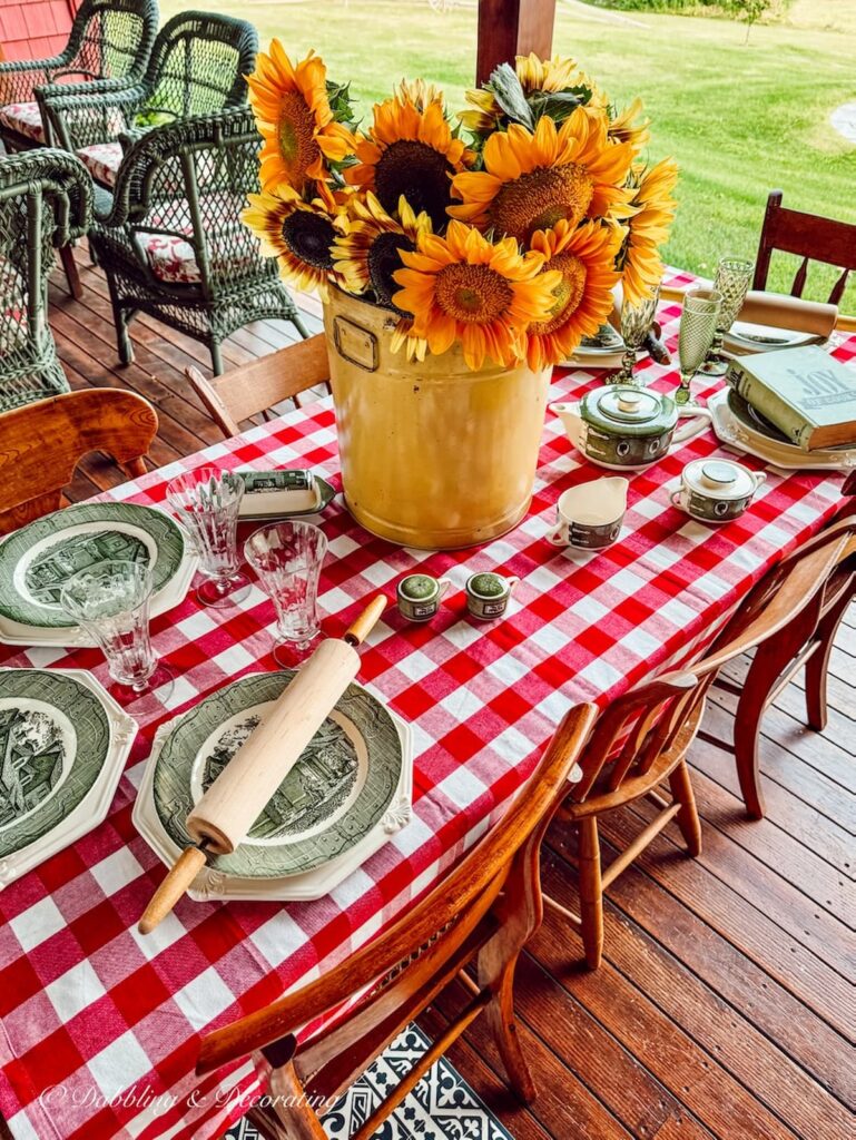 Colorful table decor with red gingham tablecloth, yellow bucket of sunflowers and Royal Colonial thrifted dishes on outdoor porch.