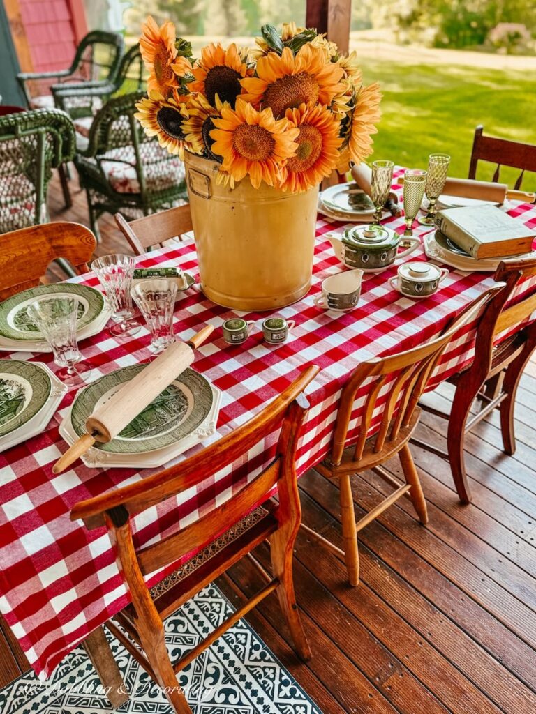 Outdoor porch table decorated with red gingham tablecloth, vintage yellow tin of sunflowers and Royal Colonial thrifted dinner place settings.