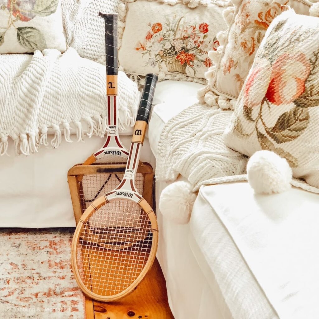 White sectional couch with vintage pillows and cable knit blankets layered in and two leaning vintage tennis rackets.