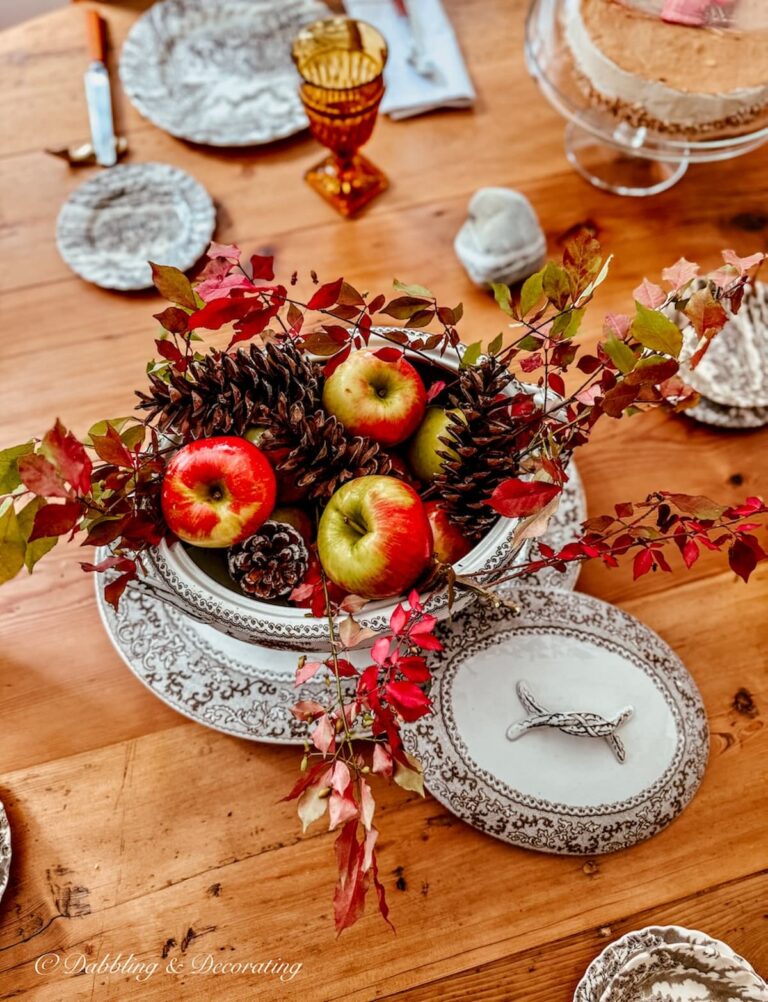 Round table centerpiece with vintage brown and white transferware soup tureen and platter filled with apples and pinecones and branches for fall decor.