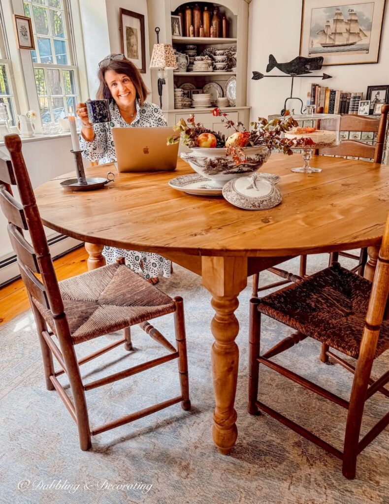 Ann, vintage home decor blogger sitting at a vintage wooden round table in a New England dining room with antique chairs.