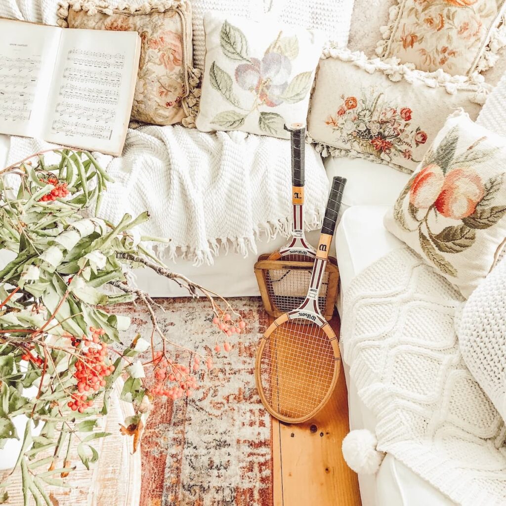 White sectional couch with vintage pillows and cable knit blankets layered in and two leaning vintage tennis rackets.