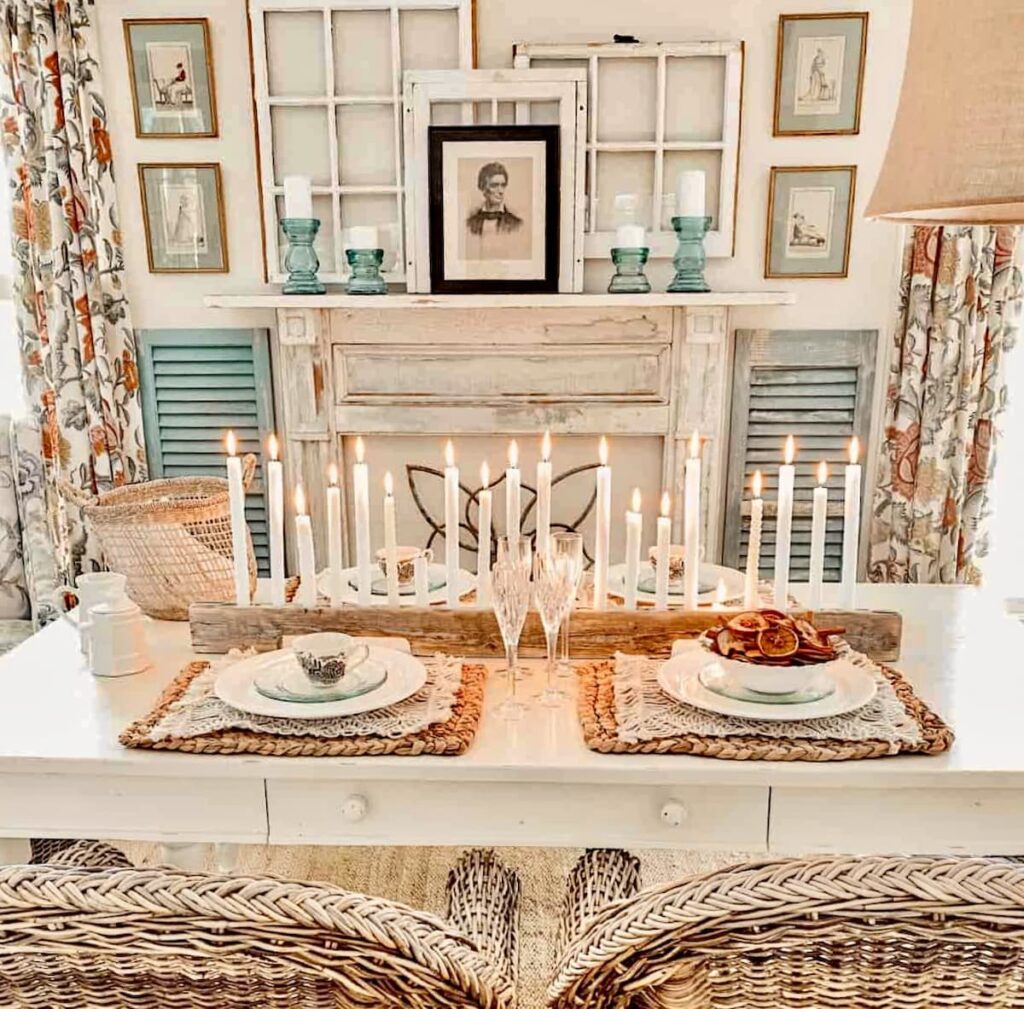 White farmhouse rectangular table decorated with candle centerpiece and faux mantel with layered old windows, shutters and wicker chairs.