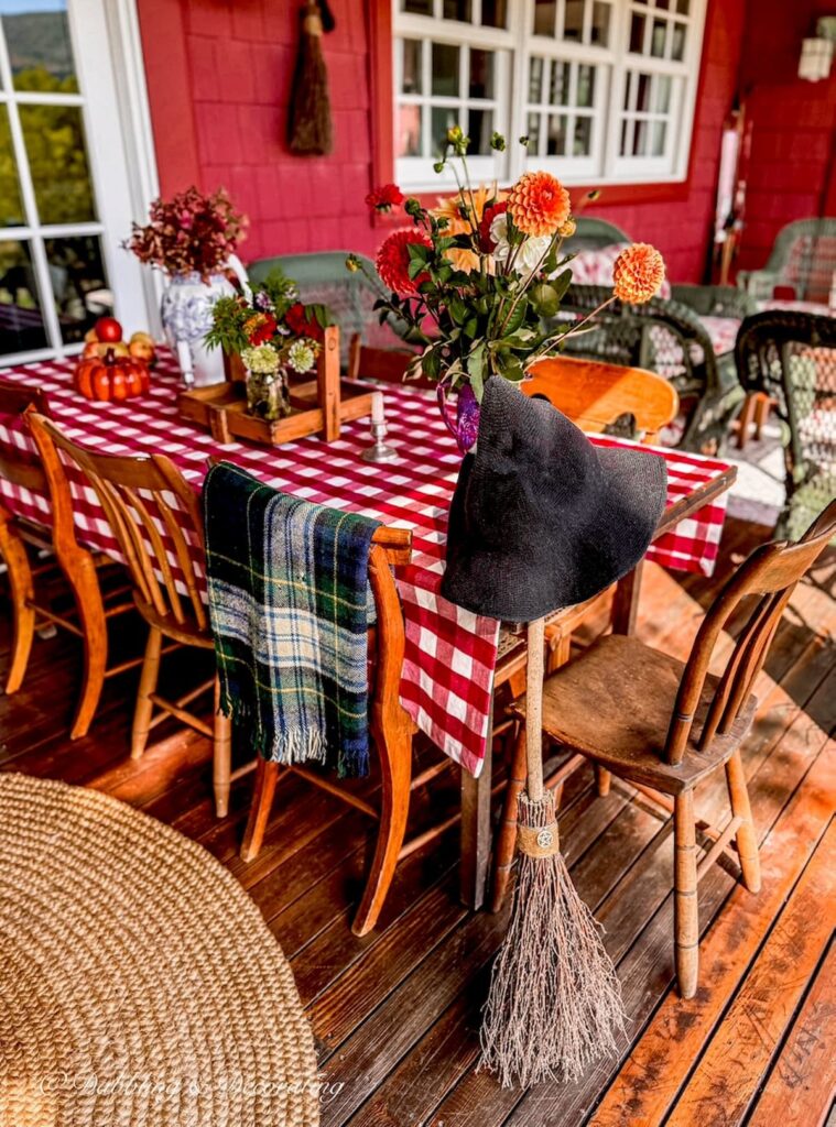 Fall porch decor with red gingham tablecloth on table and witch's broom with hat leaning on it with vintage chairs, florals, and plaid blanket.