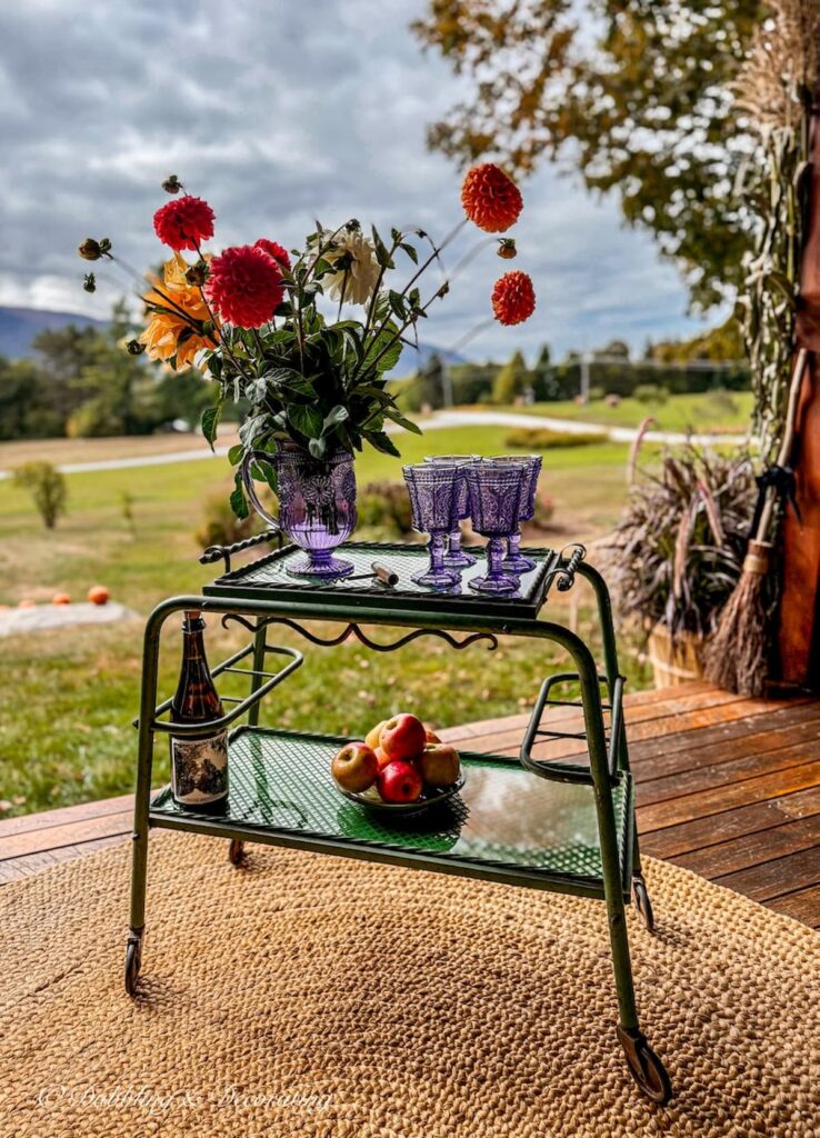 Antique beverage cart on fall porch with bouquet of fall flowers, amethyst glassware and bowl of apples with mountain views.