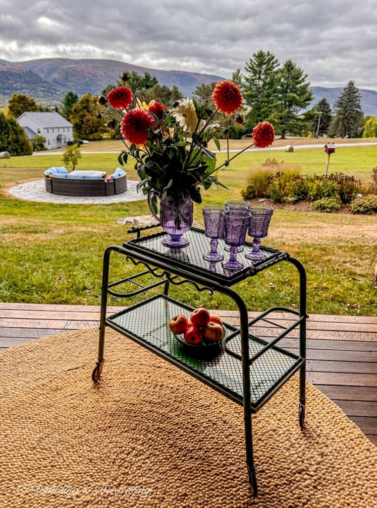Antique beverage cart on fall porch with bouquet of fall flowers, amethyst glassware and bowl of apples with mountain views.
