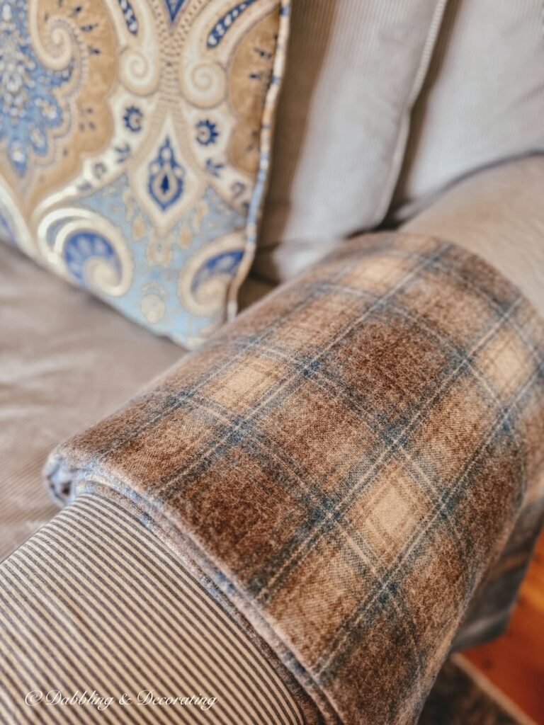 Close up of blue and brown plaid vintage Woolrich blanket size swatch folded over blue and white striped love seat armrest next to blue and brown paisley pillow in living room.