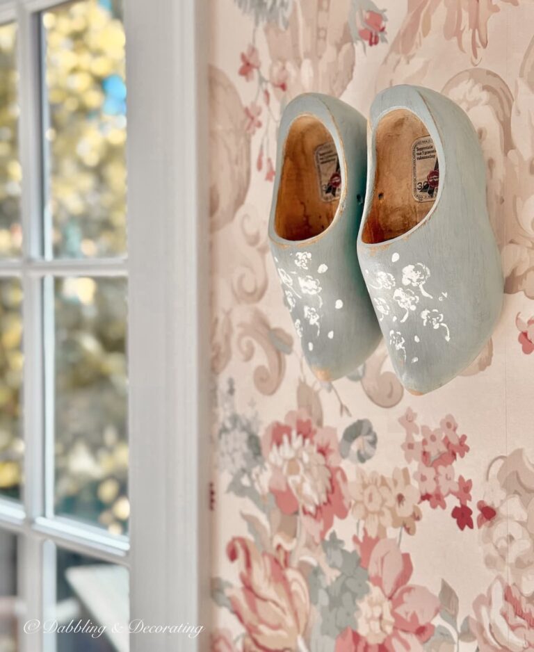 Wooden shoes painted blue with white flower stenciling hanging in wall decor on pink wallpaper in eclectic home decor project.