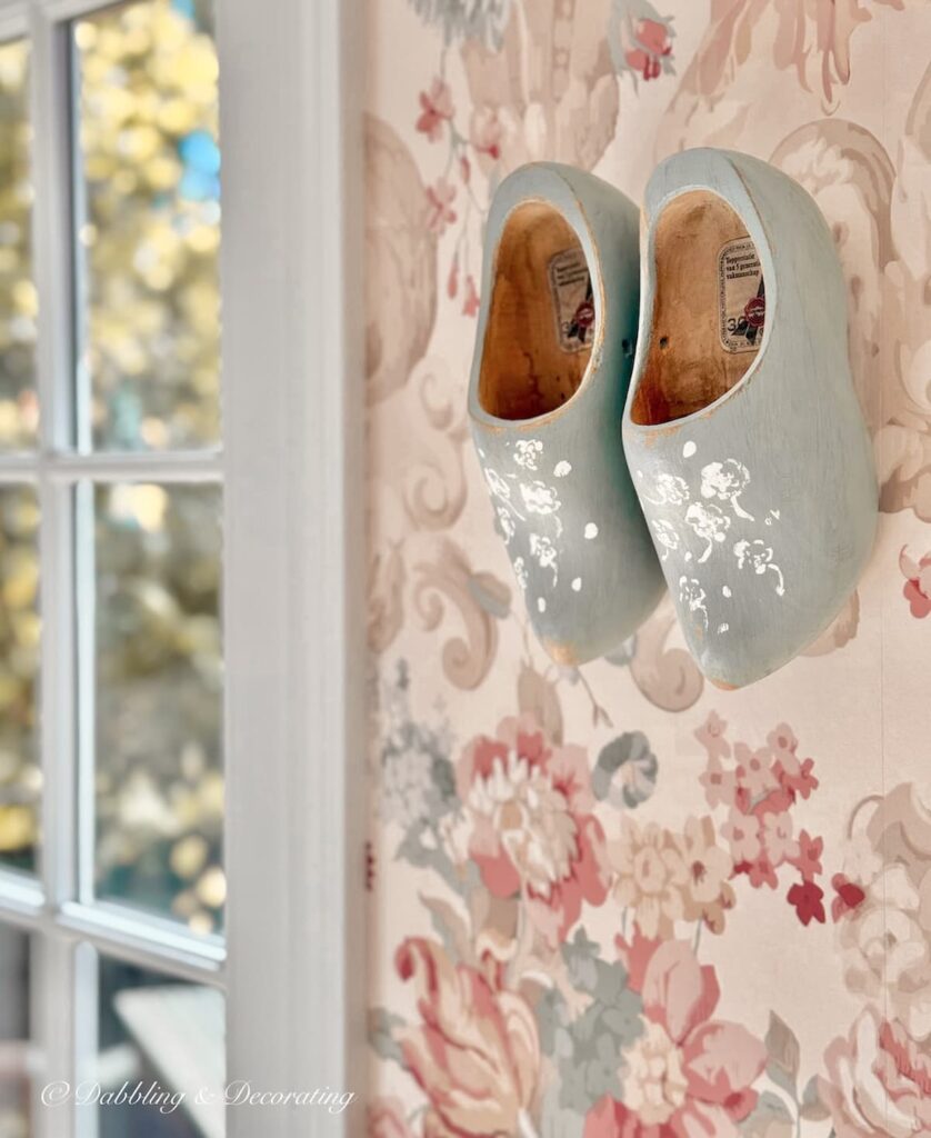 Wooden shoes painted blue with white flower stenciling hanging in wall decor on pink wallpaper in eclectic home decor project.