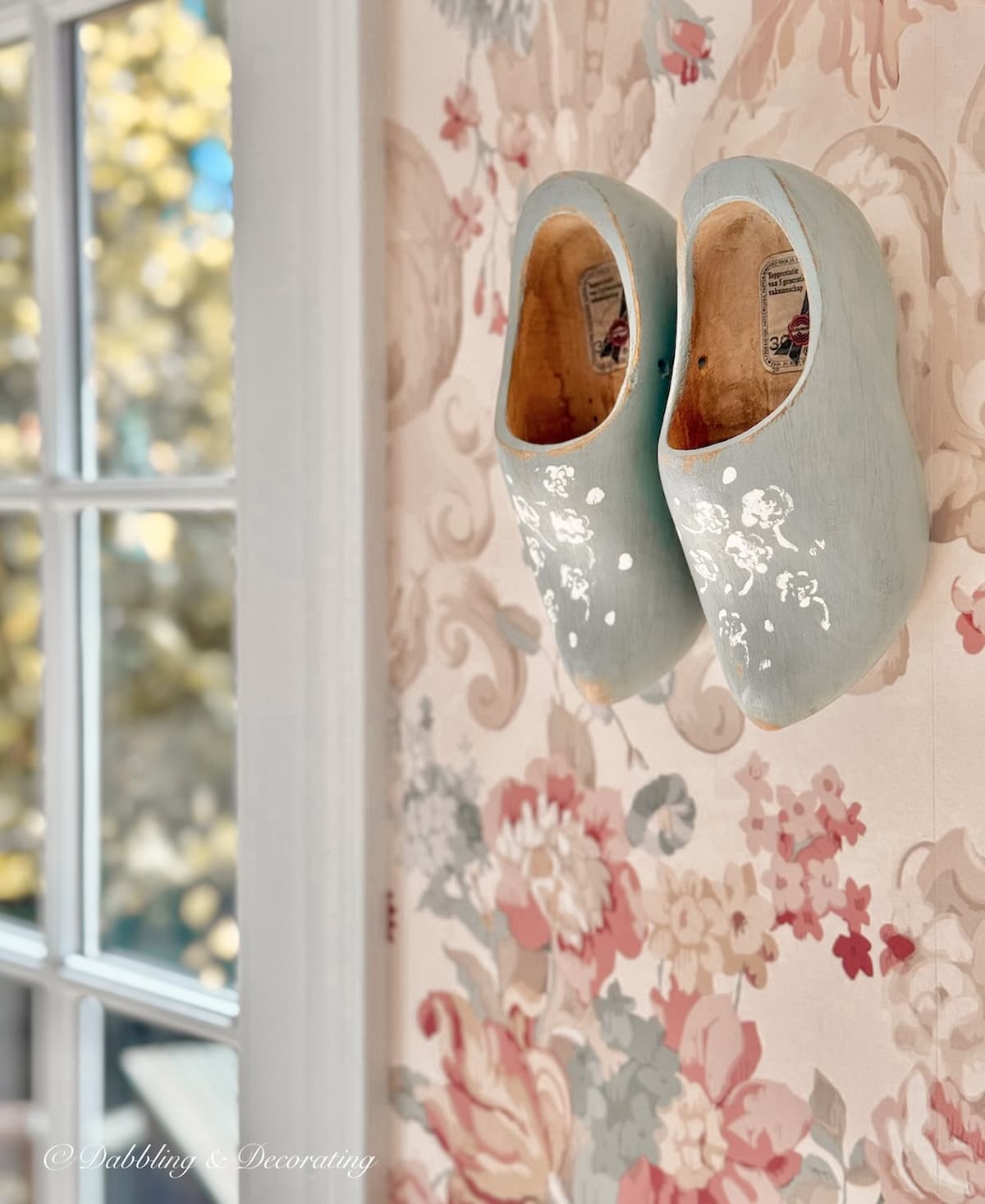 Wooden shoes painted blue with white flower stenciling hanging in wall decor on pink wallpaper in eclectic home decor project.
