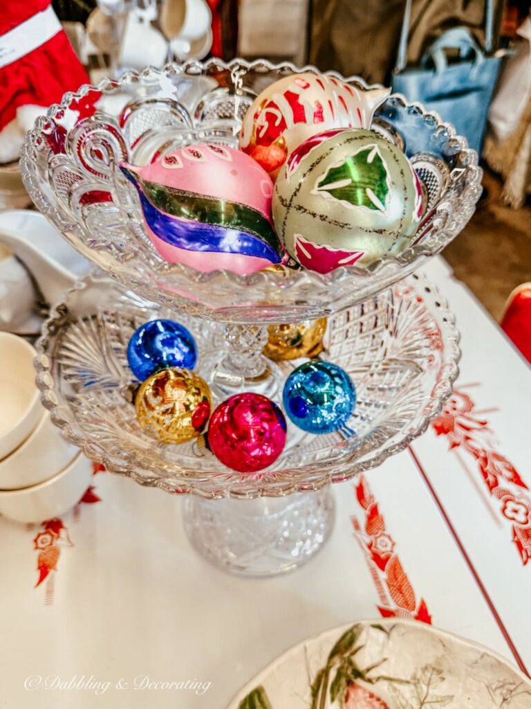 Vintage Christmas ornament bulbs on glass vintage tiered tray displayed in vintage Christmas decor at a market.