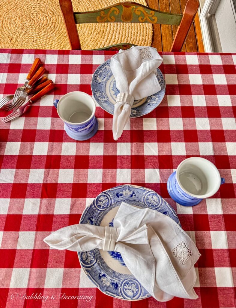 Red gingham tablecloth on porch table with blue and white vintage place settings and vintage home decor gifts, Pennsylvania Dutch style painted cane chairs.