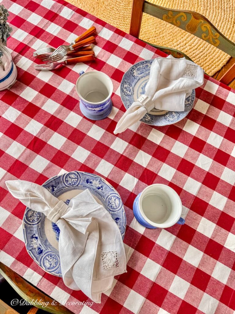 Red gingham tablecloth on porch table with blue and white vintage place settings and vintage home decor gifts, Pennsylvania Dutch style painted cane chairs.