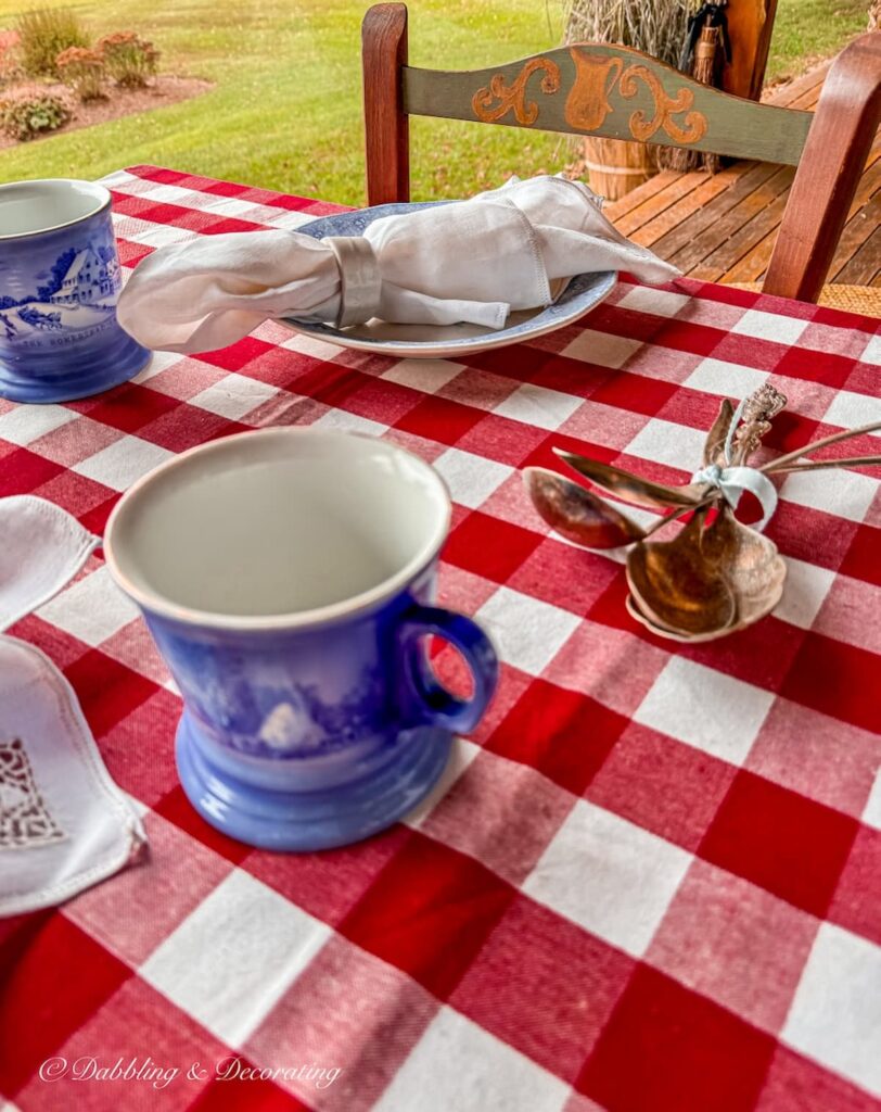 Red gingham tablecloth on porch table with blue and white vintage place settings and vintage home decor gifts, Pennsylvania Dutch style painted cane chairs.
