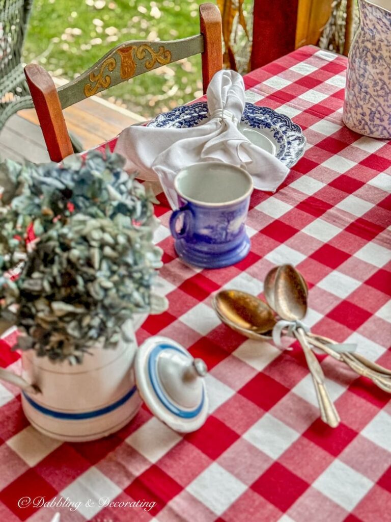 Red gingham tablecloth on outdoor porch table with blue and white place settings and Pennsylvania Dutch Style chairs.