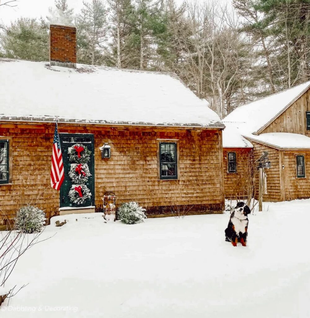 Cedar shake home in the snow with three Christmas wreaths with red bows on front door with Bernese Mountain Dog.