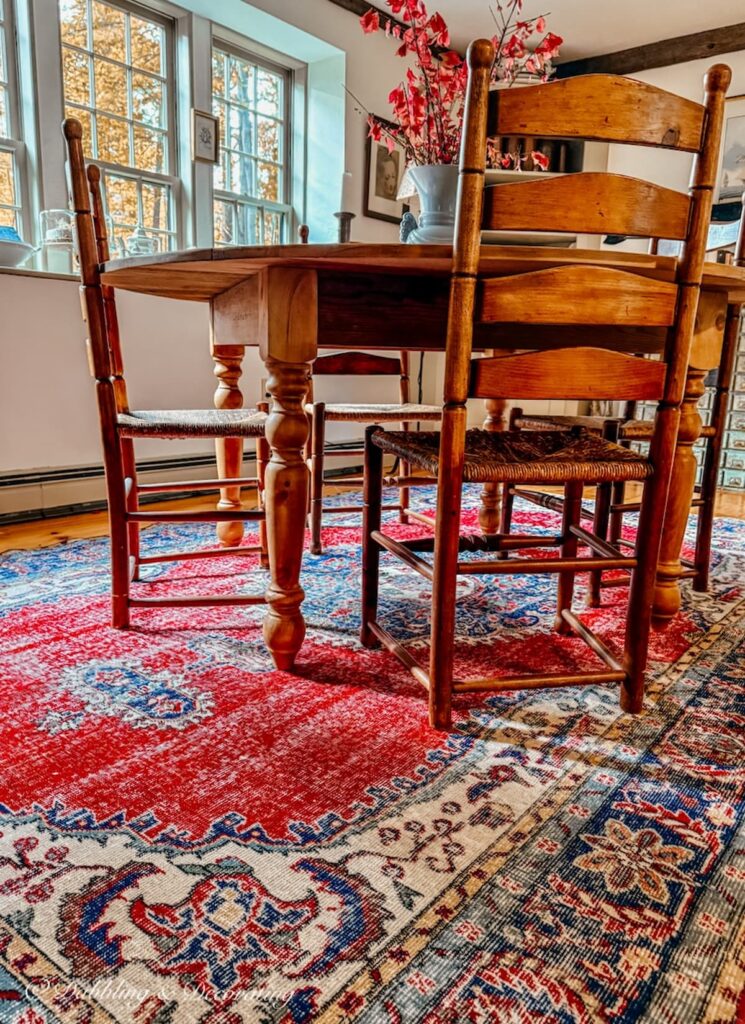 Vintage Turkish red rug in dining room with round table and wooden ladder back chairs on soft pine flooring in antique style room.