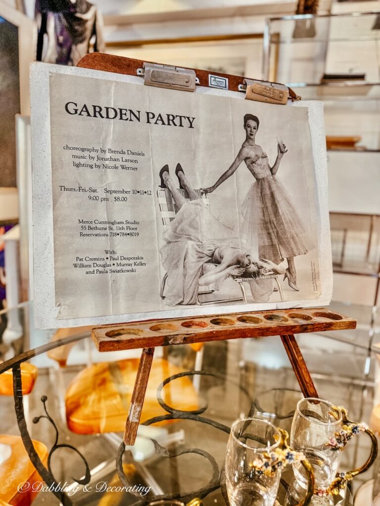 Small vintage painter's easel with Garden Party NYC play advertisement with vintage glassware on table at The Antique Warehouse Hudson, NY