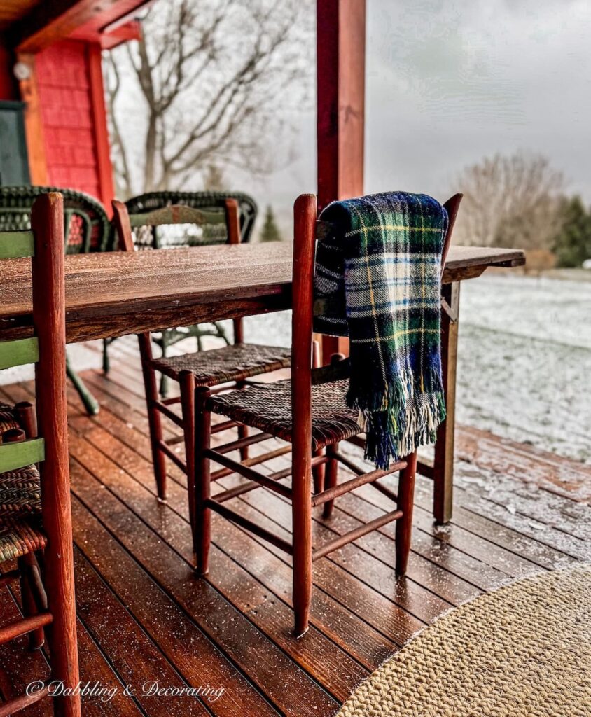Outdoor wooden porch table with chairs and thrifted plaid blanket draped over chair with light snow in Vermont.