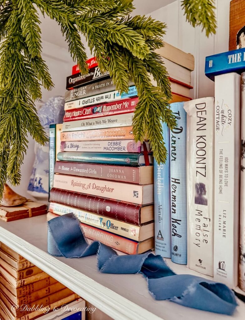 Christmas book shelf decor with stacked piles of eclectic books and a resting blue satin ribbon with evergreens.