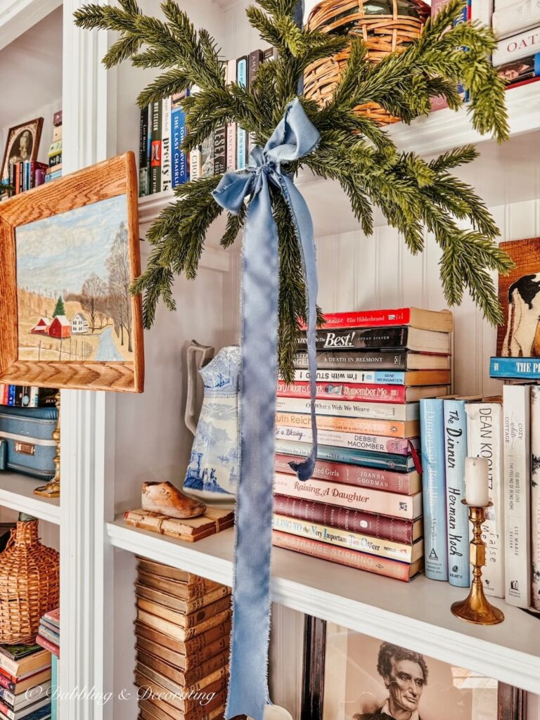 Eclectic Christmas Book shelf Decor with Faux evergreen snowflakes, blue ribbon, stacks of books and framed wooden country scene layered in.
