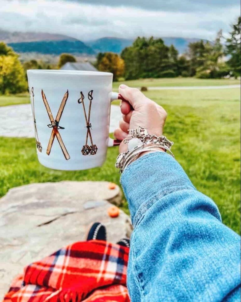 Woman holding up a vintage ski mug collection with red plaid blanket to the mountains of Vermont off of winter decor style porch.