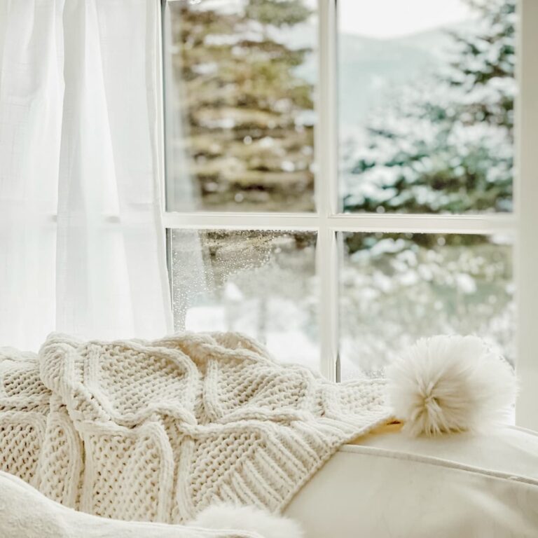 Soft white knit blanket and pom pom hat on a cream couch, with snow outside the window—a winter white decor moment.