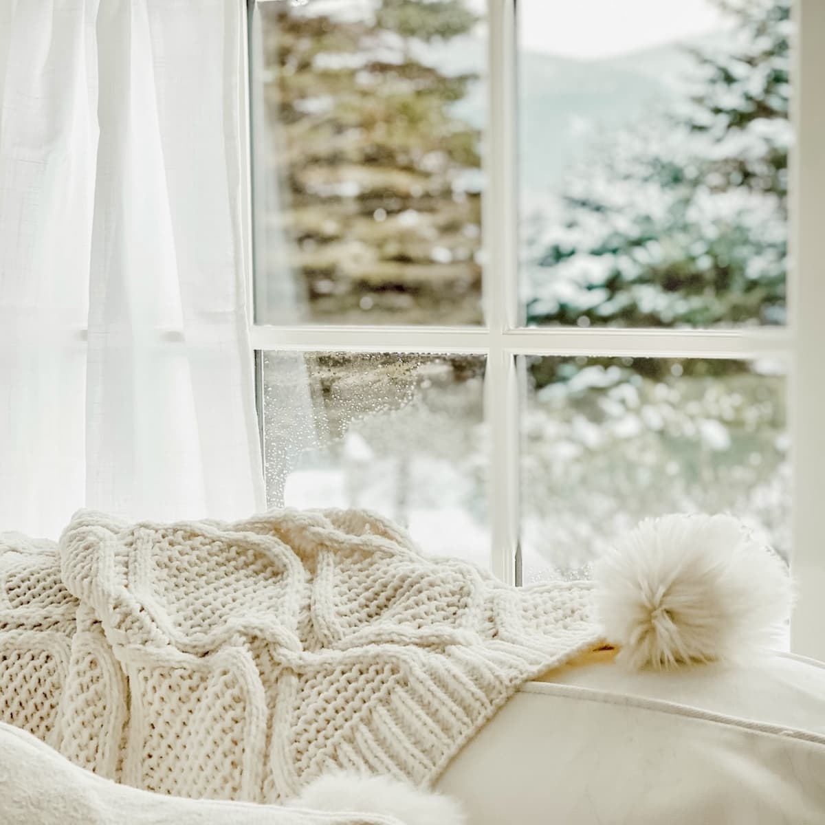 Soft white knit blanket and pom pom hat on a cream couch, with snow outside the window—a winter white decor moment.