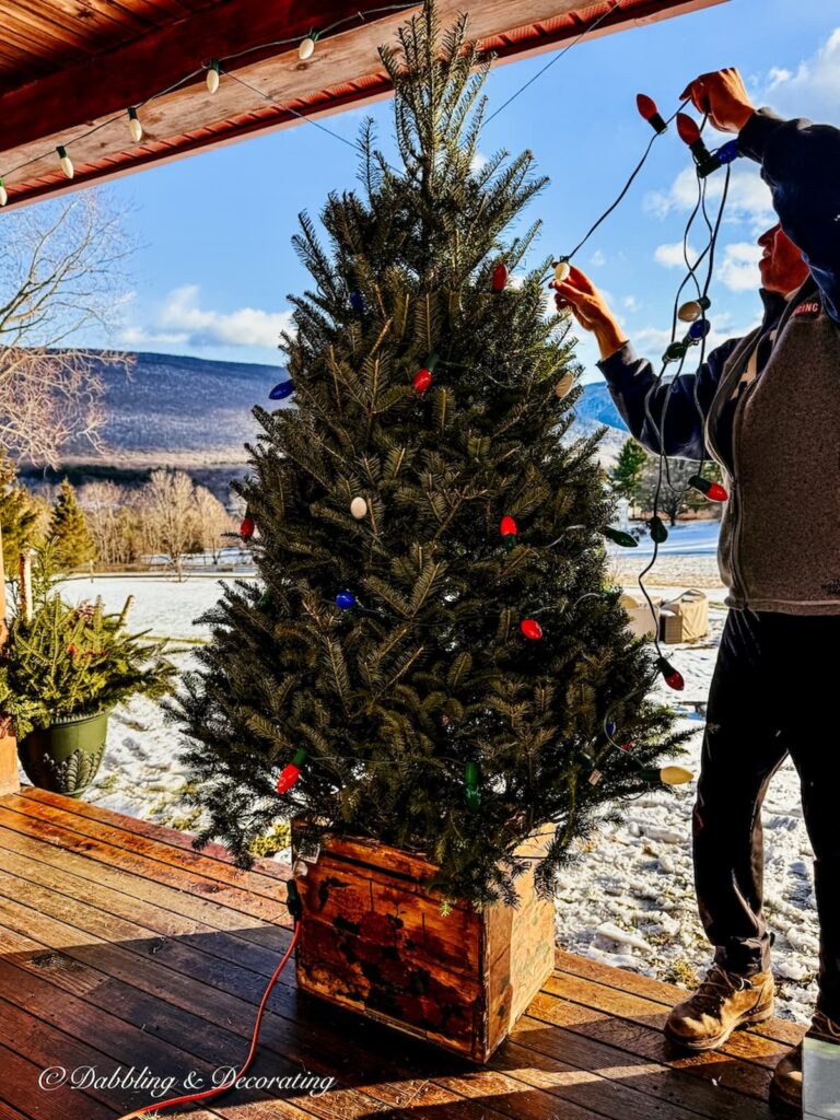 Man decorating an apres ski Christmas decor on a front porch of a Vermont home with retro Christmas lights and an old crate.