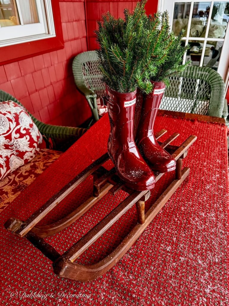 Red wellie boots filled with evergreens on small sled on Après ski Christmas porch decor on table.