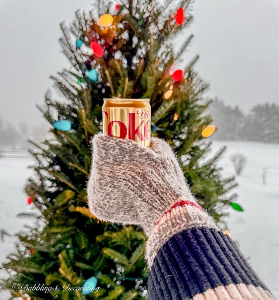 Mug mitten with diet coke in hand toasting to Après Ski Christmas Porch Decor Christmas tree in the snow.