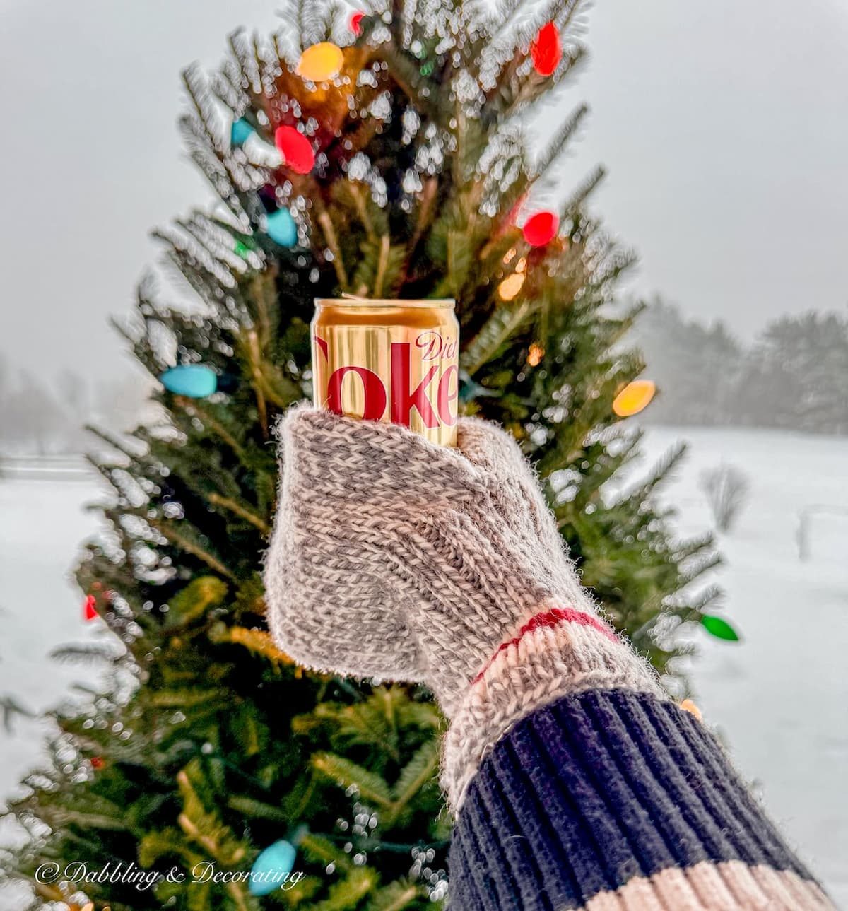Mug mitten with diet coke in hand toasting to Après Ski Christmas Porch Decor Christmas tree in the snow.