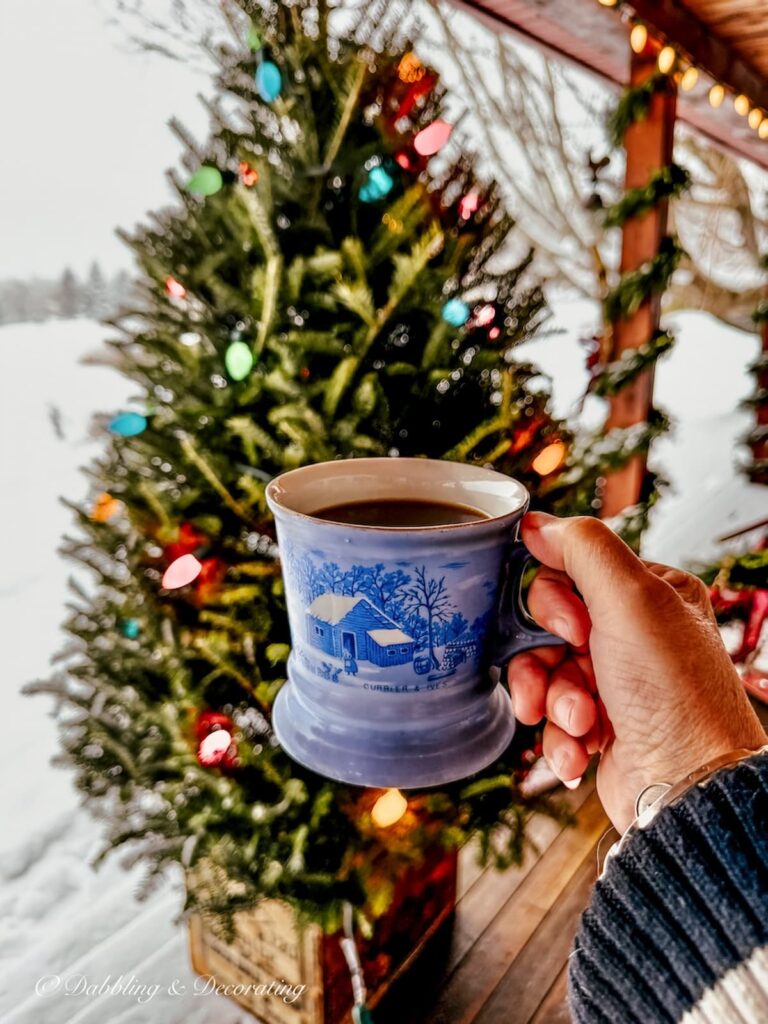 Après Ski Christmas Porch Decor with coffee in a blue vintage mug in hand cheering to a retro styled outdoor Christmas tree in a crate on a snowy Vermont day.