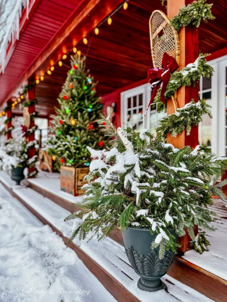 Après Ski Christmas Porch Decor on outdoor red house porch with planters filled with evergreens and retro-inspired Christmas tree in a vintage crate.
