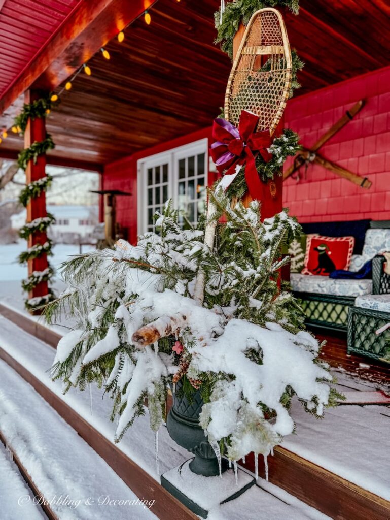 Vintage wooden snowshoe on outdoor porch beam with Christmas garland and a planter iced over with snow filled with evergreens on Vermont home.