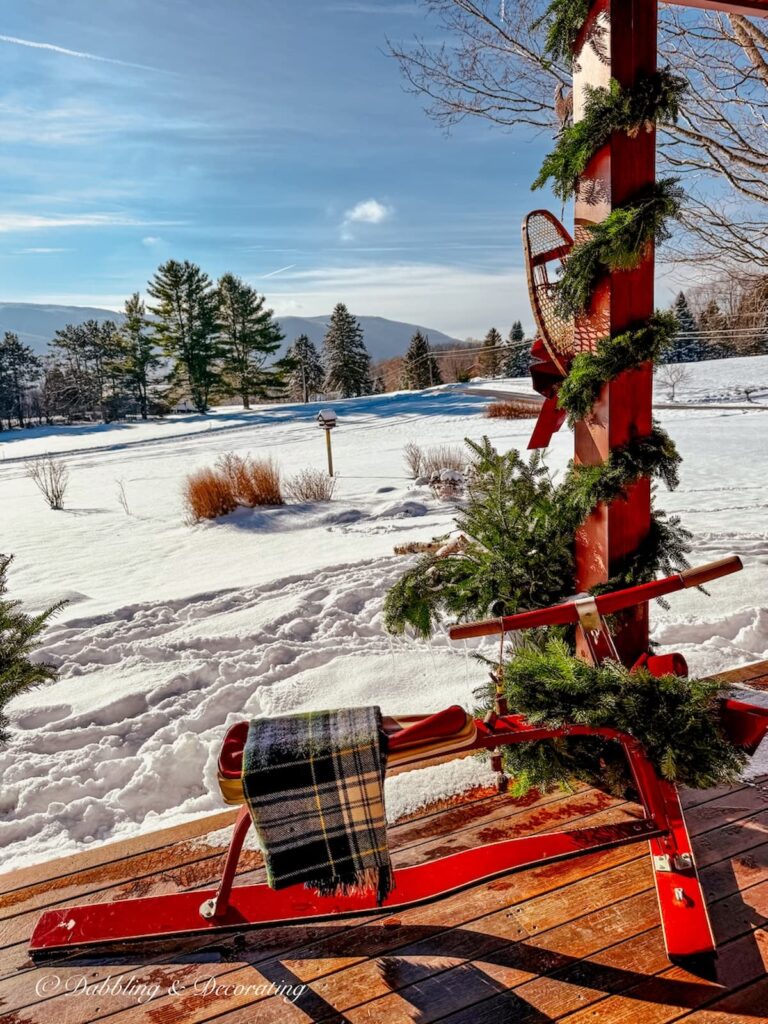 Vintage French Alps ski bob in Après Ski Christmas Porch decor in the snow with wreath and plaid blanket with mountain views and blue skies in the snow.