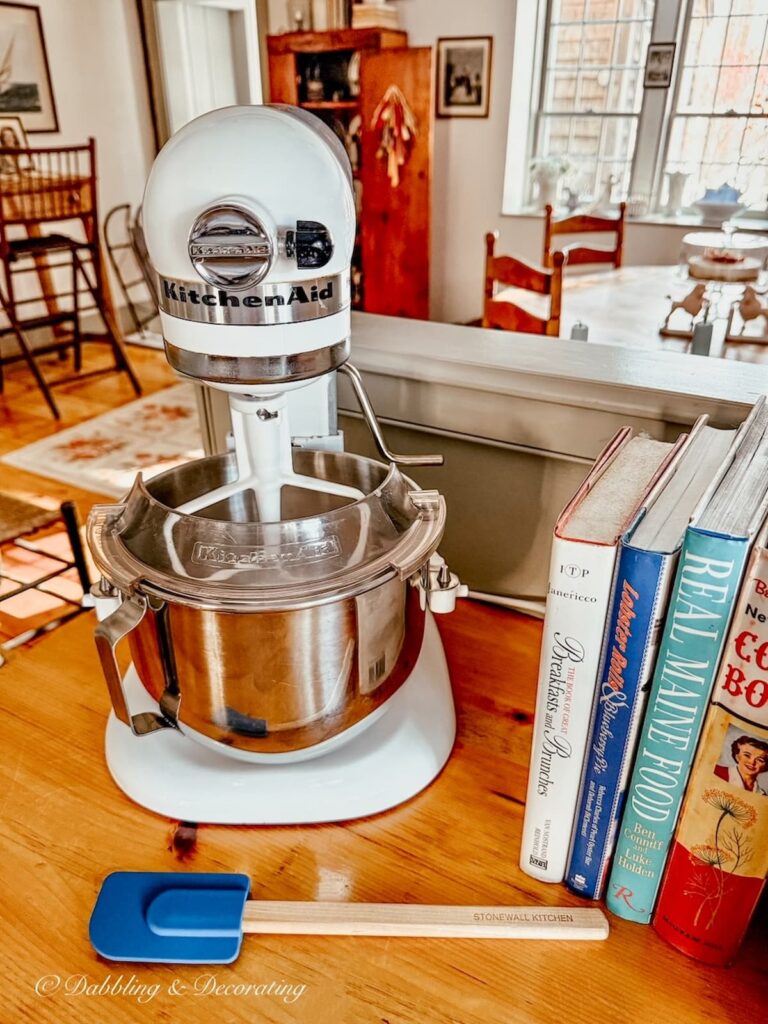 White KitchenAid Mixer on wooden kitchen countertop with blue spatula, and vintage books in thrifted winter home decor ideas.