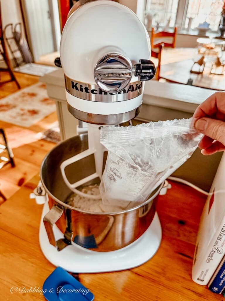 White KitchenAid Mixer with hand pouring in mix to the bowl making blueberry muffins in cozy winter home decor ideas.