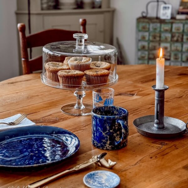 Glass cake stand filled with homemade blueberry muffins and blue pottery place setting on antique dining room table for cozy winter home decor ideas created with thrifted Goodwill Northern New England finds.
