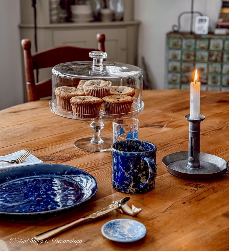 Glass cake stand filled with homemade blueberry muffins and blue pottery place setting on antique dining room table for cozy winter home decor ideas created with thrifted Goodwill Northern New England finds.