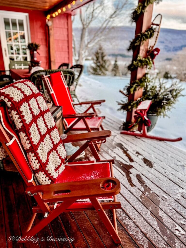 Two red folding chairs on covered porch in the snow with thrifted red and white afghan.