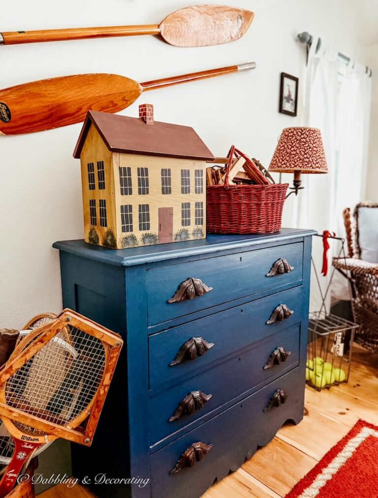 Vintage painted blue dresser in bedroom styled with vintage collectibles like vintage wooden house, tennis racquets and old boat oars on wall.
