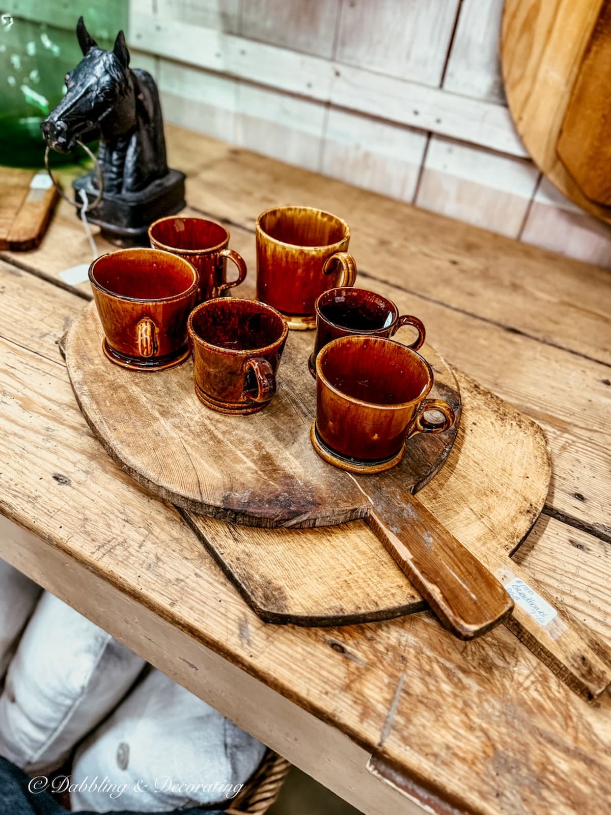 Stack of breadboards in antique store with set of brown mugs and wrought iron black horse head.